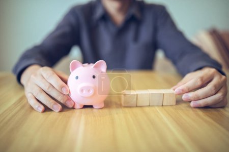 A person with a piggy bank and wooden blocks on a table, illustrating concepts of savings and investment.