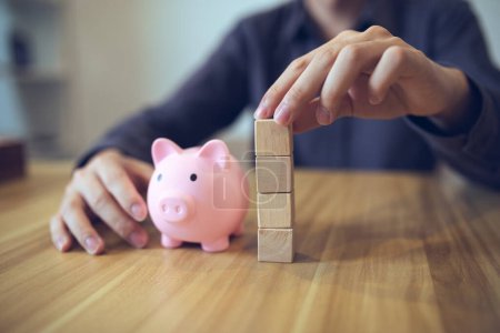 Person building a tower from wooden blocks beside a piggy bank, symbolizing financial planning and saving.