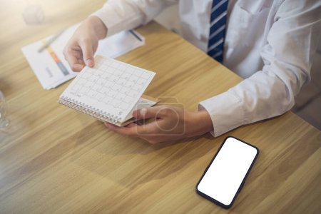 Professional in a white shirt scheduling in a diary with a smartphone with screen mockup on the desk for quick access.
