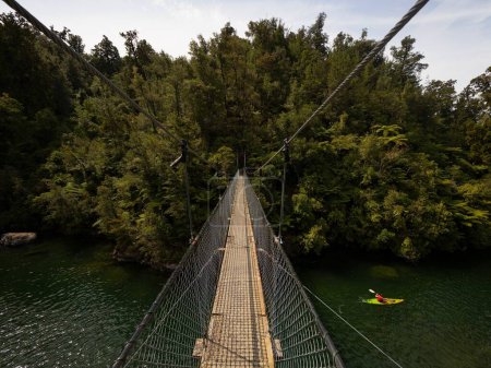 Téléchargez les photos : Swinging cable suspension foot bridge spanning over river fresh water stream with person in kayak in Abel Tasman National Park South Island New Zealand - en image libre de droit