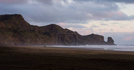 A couple walking on idyllic black sand Te Henga Bethells beach with rock formation in background during sunset in West Auckland North Island New Zealand