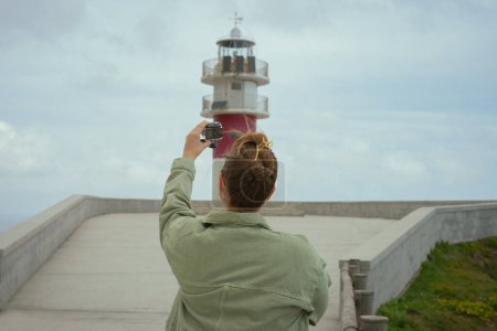Young woman filming herself in front of a lighthouse on a cloudy day. Summer vacation concept.
