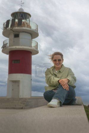 Young woman with sunglasses sitting posing in front of the lighthouse at Cabo Ortegal, Galicia. Vacations in northern Spain
