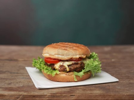 Photo for Big Burger lies on craft white paper against wooden table. A Juicy green Salad leaf and a red Tomato lie near the Burger - Royalty Free Image