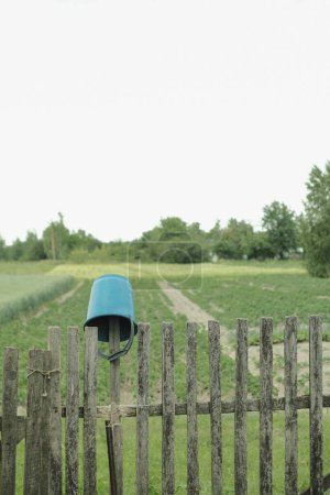 Téléchargez les photos : Un seau vert est accroché à une vieille clôture pourrie du village sur fond de potager. - en image libre de droit