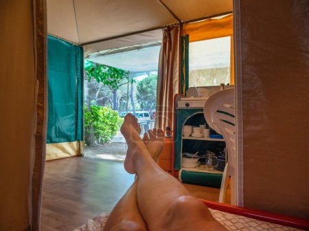 Photo for Legs of a girl lying on the bed of a camping tent. View of the kitchen and the terrace. Vacation concept. - Royalty Free Image