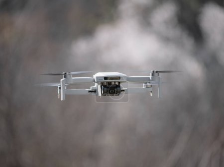 Petit drone blanc avec une caméra intégrée à mi-vol et un fond flou d'arbres aux fleurs blanches donnant la sensation de fumée autour et la vitesse en général
