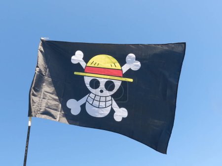 Bandera pirata negra que contiene un cráneo blanco con un sombrero de paja amarillo y una cinta roja ubicada en dos huesos blancos, ondeando en el viento sobre un mástil y el cielo azul en el fondo
