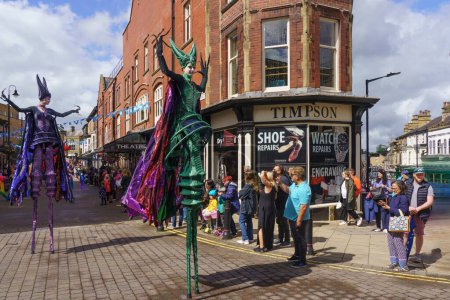 Téléchargez les photos : The Carnival Parade in Harrogate, mettant en vedette deux danseuses de Stilt en costumes verts et violets avec de longues manteaux marchant à travers le centre-ville, North Yorkshire, Angleterre, Royaume-Uni. - en image libre de droit