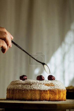 Téléchargez les photos : Main d'homme avec cuillère finissant la préparation du gâteau sucré fait maison, saupoudré de sucre en poudre avec des cerises - en image libre de droit