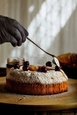 Téléchargez les photos : Main d'homme avec cuillère finissant la préparation du gâteau sucré fait maison, saupoudré de sucre en poudre avec des cerises - en image libre de droit