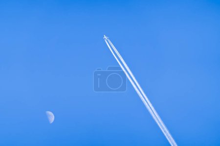 Ein Flugzeug, das durch den azurblauen Himmel fliegt und weiße Kondensstreifen hinterlässt, mit dem Mond an der Seite, inmitten des Nachmittags.