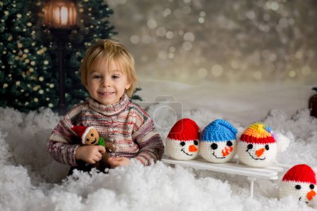 Téléchargez les photos : Enfant tout-petit mignon, tenant un jouet tricoté, jouant dans la neige avec des bonhommes de neige colorés, souriant joyeusement - en image libre de droit