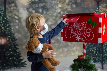 Téléchargez les photos : Petit tout-petit mignon, doux garçon blond, envoi d'une lettre au Père Noël, portant un masque médical et tenant un ours en peluche, coronavirus, Noël 2020 - en image libre de droit