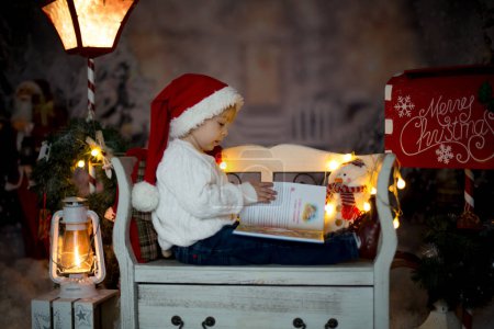 Foto de Hermoso niño, niño rubio, leyendo un libro, sentado en un banco con decoración navideña a su alrededor - Imagen libre de derechos