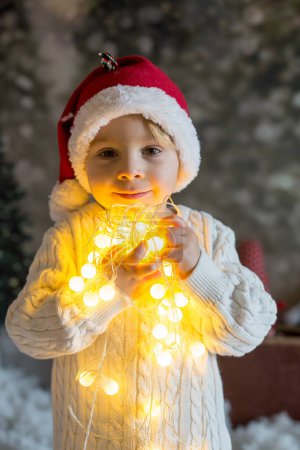Foto de Hermoso niño pequeño, sosteniendo la cadena ligera de Navidad, mirando a la cámara, retrato de Navidad cercano - Imagen libre de derechos
