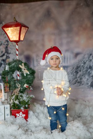 Foto de Lindo niño pequeño, niño rubio, de pie frente a la casa, envuelto en luces de cuerda de Navidad, sintiéndose atrapado - Imagen libre de derechos