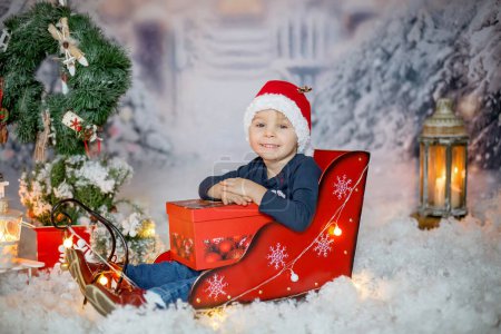 Foto de Lindo niño rubio pequeño, niño, sentado en trineo al aire libre, escena de invierno nevado, decoración de Navidad a su alrededor - Imagen libre de derechos