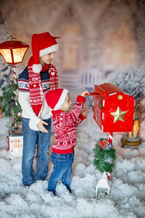 Photo for Cute children, school boy and toddler, sending letter to santa in christmas mailbox, christmas decoration around him, outdoor shot, outdoor snow shot - Royalty Free Image