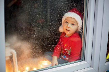 Foto de Hermoso niño pequeño, muchacho, esperando en la ventana en la víspera de Navidad, en busca de Santa Claus curiosamente - Imagen libre de derechos