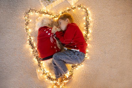 Photo for Cute blond boy and his pet dog in sweater, lying in a heart from light string on the floor, smiling at camera - Royalty Free Image