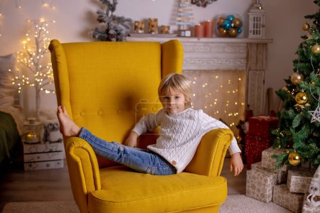 Téléchargez les photos : Mignon enfant, garçon, assis dans un fauteuil jaune dans une chambre décorée pour Noël avec la mère et la grand-mère - en image libre de droit