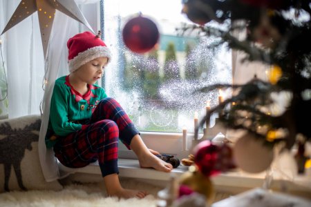 Photo for Toddler child, cute blond boy, sitting on the window in pajama, looking out for Satna Claus,, eating cookies,  Christmas lights around him - Royalty Free Image