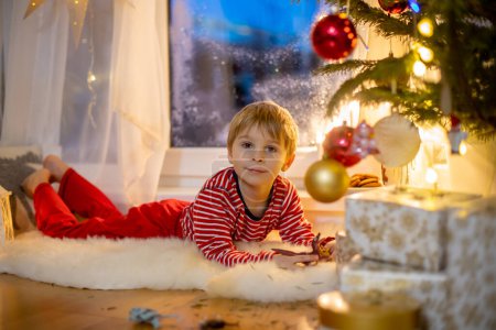 Foto de Toddler child, cute blond boy, sitting on the window in pajama, looking out for Satna Claus,, eating cookies,  Christmas lights around him - Imagen libre de derechos