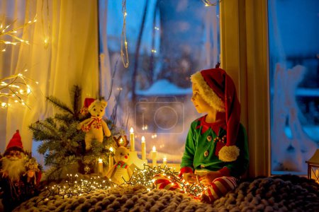 Foto de Toddler child, cute blond boy, sitting on the window, reading book, Christmas lights around him - Imagen libre de derechos