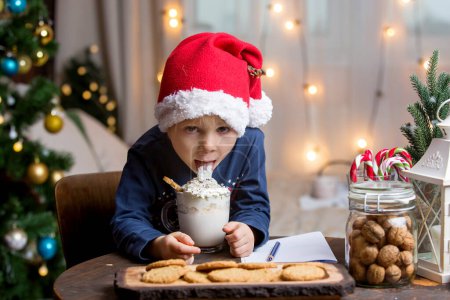 Téléchargez les photos : Beau blond tout-petit enfant, garçon, boire du chocolat chaud avec crème essuyée, décoration de Noël autour de lui - en image libre de droit