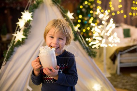 Foto de Hermoso niño rubio, niño, beber cacao caliente con crema limpiada, decoración de Navidad a su alrededor - Imagen libre de derechos