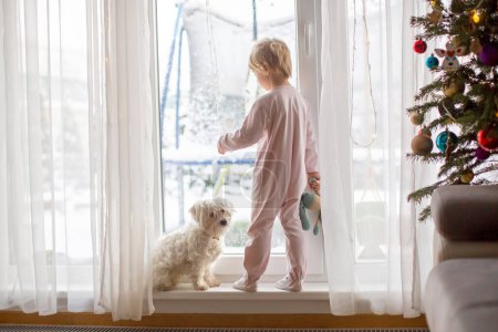 Foto de Lindo niño pequeño en pijama, de pie frente a unas grandes ventanas francesas con su perro mascota, disfrutando de la nieve afuera - Imagen libre de derechos
