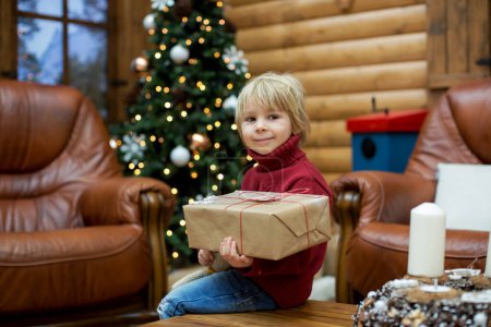 Téléchargez les photos : Enfant tout-petit blond, livre de lecture et tenue de cadeaux dans un chalet confortable dans les montagnes, période de Noël - en image libre de droit