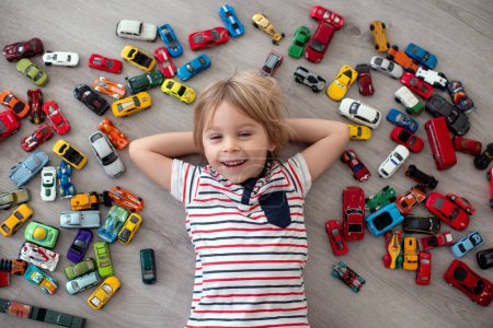 Téléchargez les photos : Enfant tout-petit mignon, garçon blond, jouer avec des voitures colofrul, différentes tailles et couleurs sur le sol, souriant - en image libre de droit