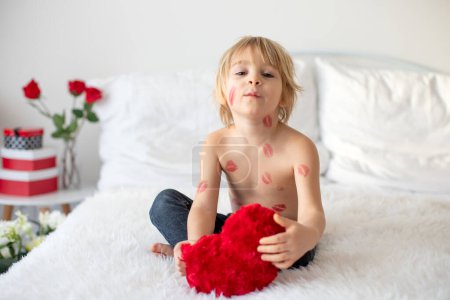 Foto de Hermoso niño rubio, niño con besos de lápiz labial en su cuerpo, sosteniendo rosa roja para San Valentín, sentado en la cama - Imagen libre de derechos