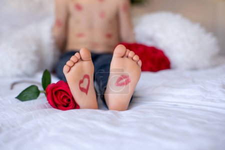 Photo for Beautiful blond toddler child, boy with lipstick kisses on his body, holding red rose for Valentine, sitting in bed - Royalty Free Image