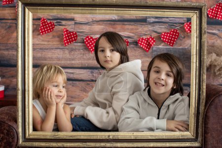 Photo for Cute toddler blond child and older brothers with valentine decoration, love, flowers, sitting on armchair with frame in studio - Royalty Free Image