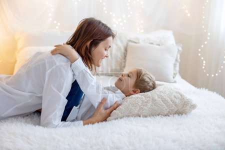 Foto de Cute blonde toddler child, boy, cuddling  in mother, sitting on the bed, pure white background with yellow soft lights - Imagen libre de derechos