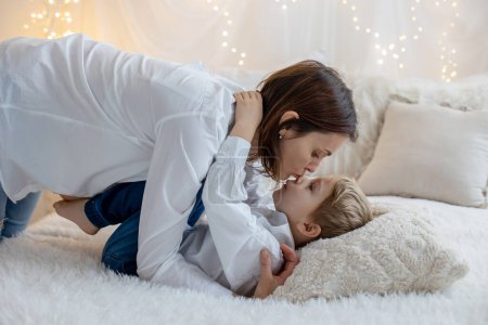 Foto de Cute blonde toddler child, boy, cuddling  in mother, sitting on the bed, pure white background with yellow soft lights - Imagen libre de derechos