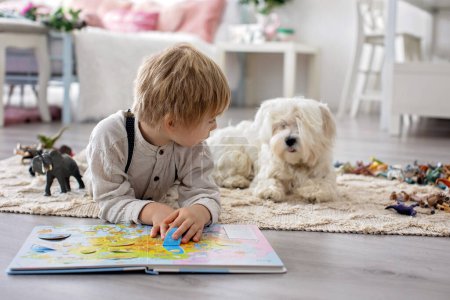 Téléchargez les photos : Mignon enfant d'âge préscolaire blond, garçon blond avec chien maltais animal de compagnie, livre de lecture à la maison sur le sol avec mère - en image libre de droit