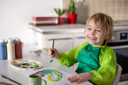 Foto de Lindo niño rubio, niño preescolar, pintura en casa con tarjeta de colores aquarelle para San Valentín - Imagen libre de derechos