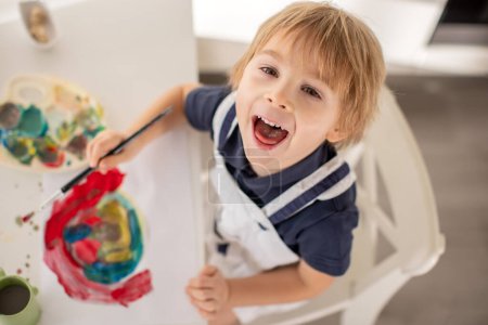 Foto de Lindo niño rubio, niño preescolar, pintura en casa con tarjeta de colores aquarelle para San Valentín - Imagen libre de derechos