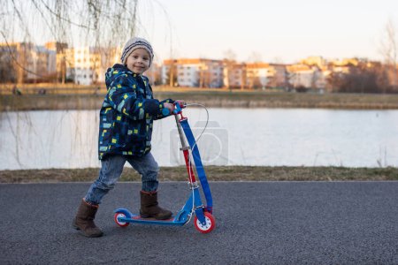 Téléchargez les photos : Mignon tout-petit garçon, équitation un scooter dans un parc sur un froid ensoleillé jour de printemps - en image libre de droit