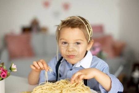 Foto de Lindo niño preescolar, chico rubio, comiendo espaguetis en casa, haciendo un desastre en todas partes, momentos divertidos - Imagen libre de derechos