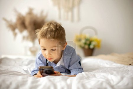 Foto de Niño rubio preescolar, lindo niño, jugando en el teléfono móvil en la cama en un día soleado - Imagen libre de derechos