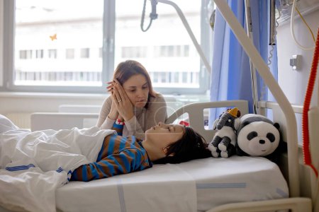 Foto de Niño preadolescente, niño, acostado en el hospital con fractura de columna torácica, vértebras, madre sentada junto a su cama - Imagen libre de derechos