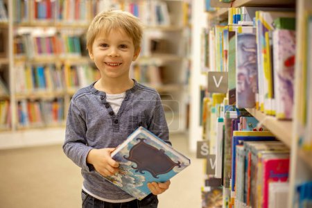 Téléchargez les photos : Adorable little child, boy, sitting in library, reading book and choosing what to lend, kid in book store - en image libre de droit