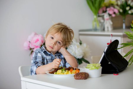 Photo for Little toddler child, blond boy, eating boiled vegetables, broccoli, potatoes and carrots with fried chicken meat at home, while watching  movie on tablet - Royalty Free Image