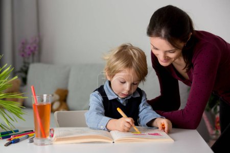 Photo for Cute preschool child, blond boy, filling some homework in a work book and coloring, mother helping him at home - Royalty Free Image