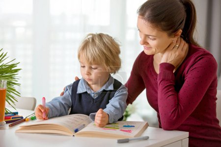 Photo for Cute preschool child, blond boy, filling some homework in a work book and coloring, mother helping him at home - Royalty Free Image